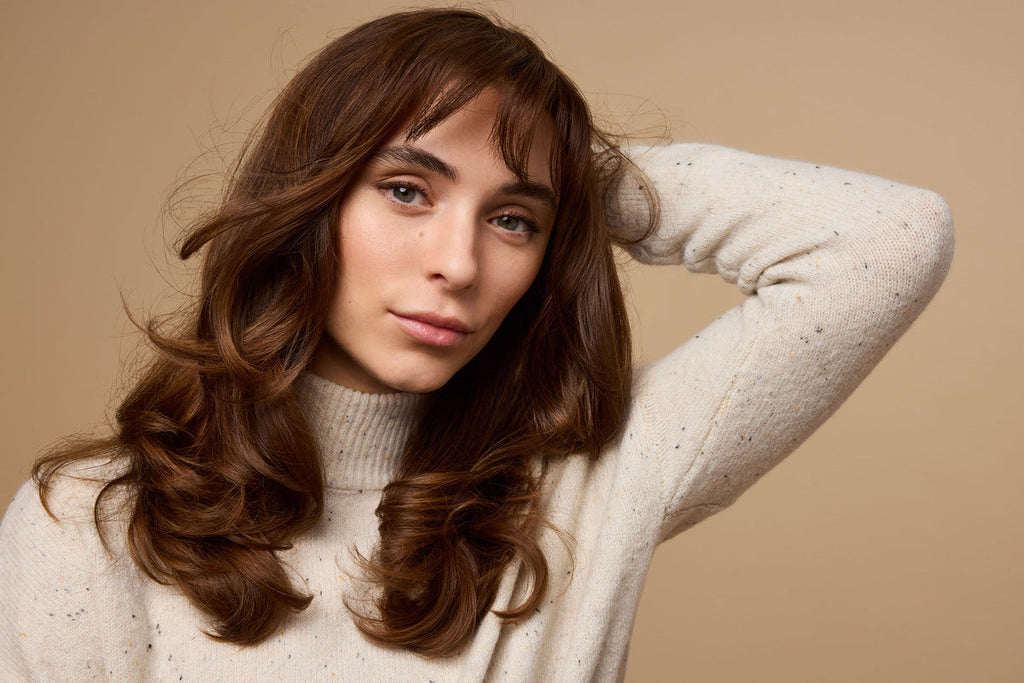 A person with long, brown hair and wearing a cream sweater poses with one hand behind their head against a warm beige background.