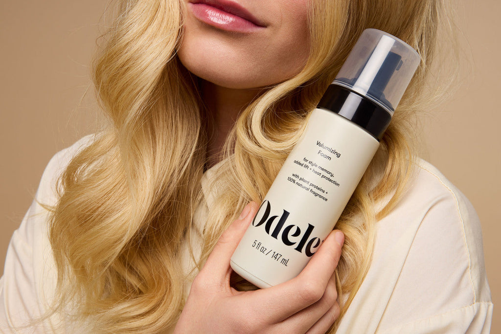 A person with fluffy blonde hair holds a bottle of Odele Volumizing Foam near their face against a beige background.