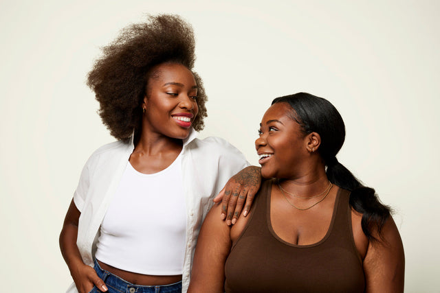 Two women, one with short, coily hair and one with long, straight hair, smile at each other.