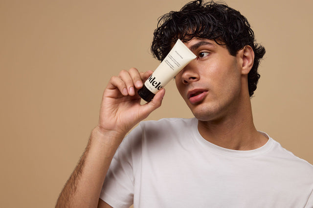 A person with short, curly brown hair holds a mini bottle of Odele Moisture Mask in front of their face