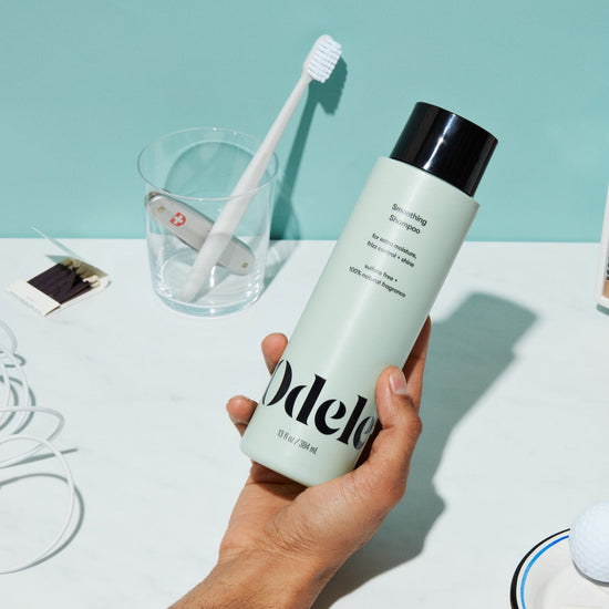 A hand holds a bottle of Odele Smoothing Shampoo over a white countertop, with a toothbrush in a clear cup, a matchbook, and tangled white earphones visible in the background.