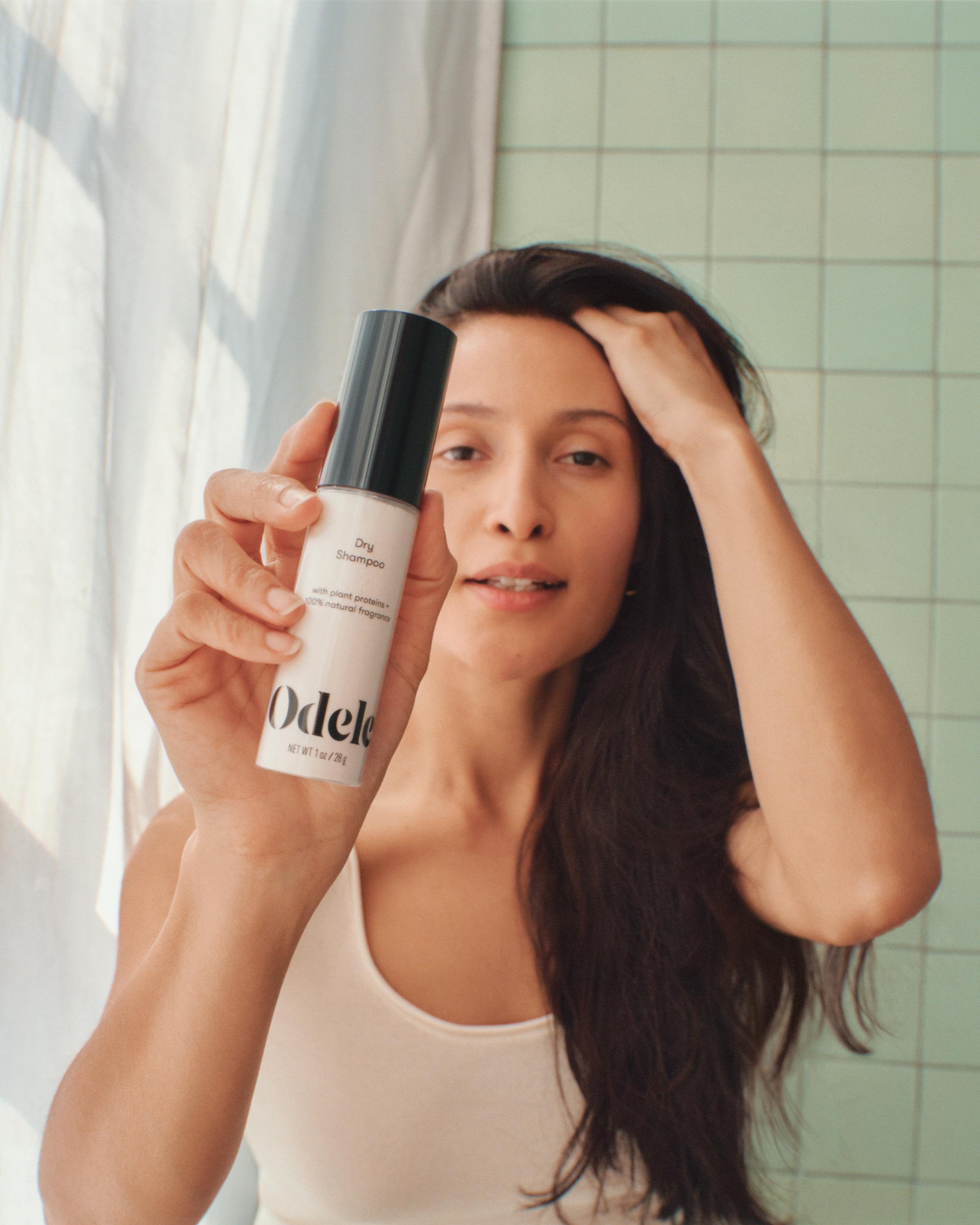 A woman with long dark hair holds up a bottle of Odele Dry Shampoo toward the camera, standing in front of a tiled wall, wearing a white tank top and touching her hair with her other hand.