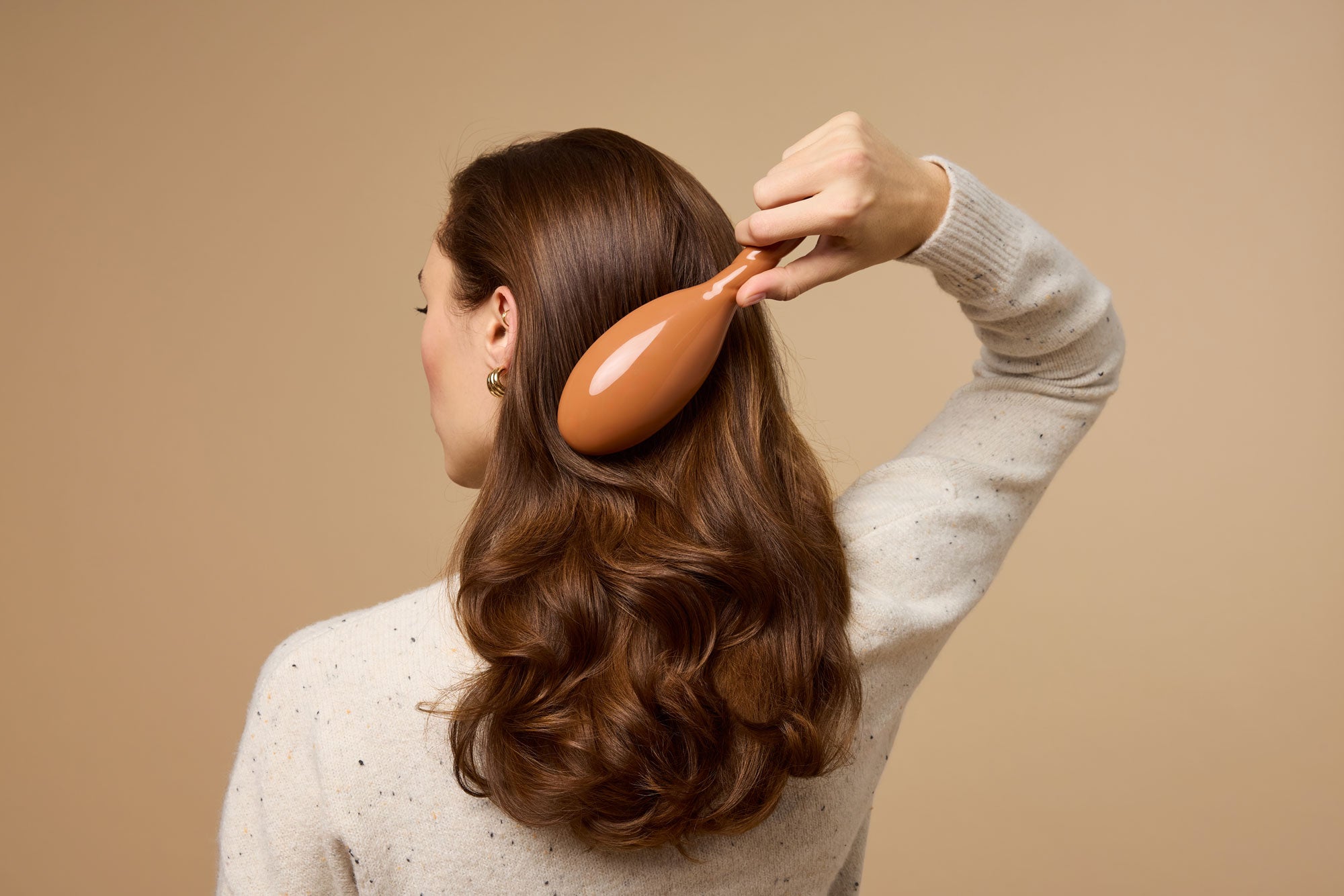 A woman with long, wavy brown hair is seen from behind, brushing her hair with a brown hairbrush. She is wearing a light-colored sweater and standing against a beige background.