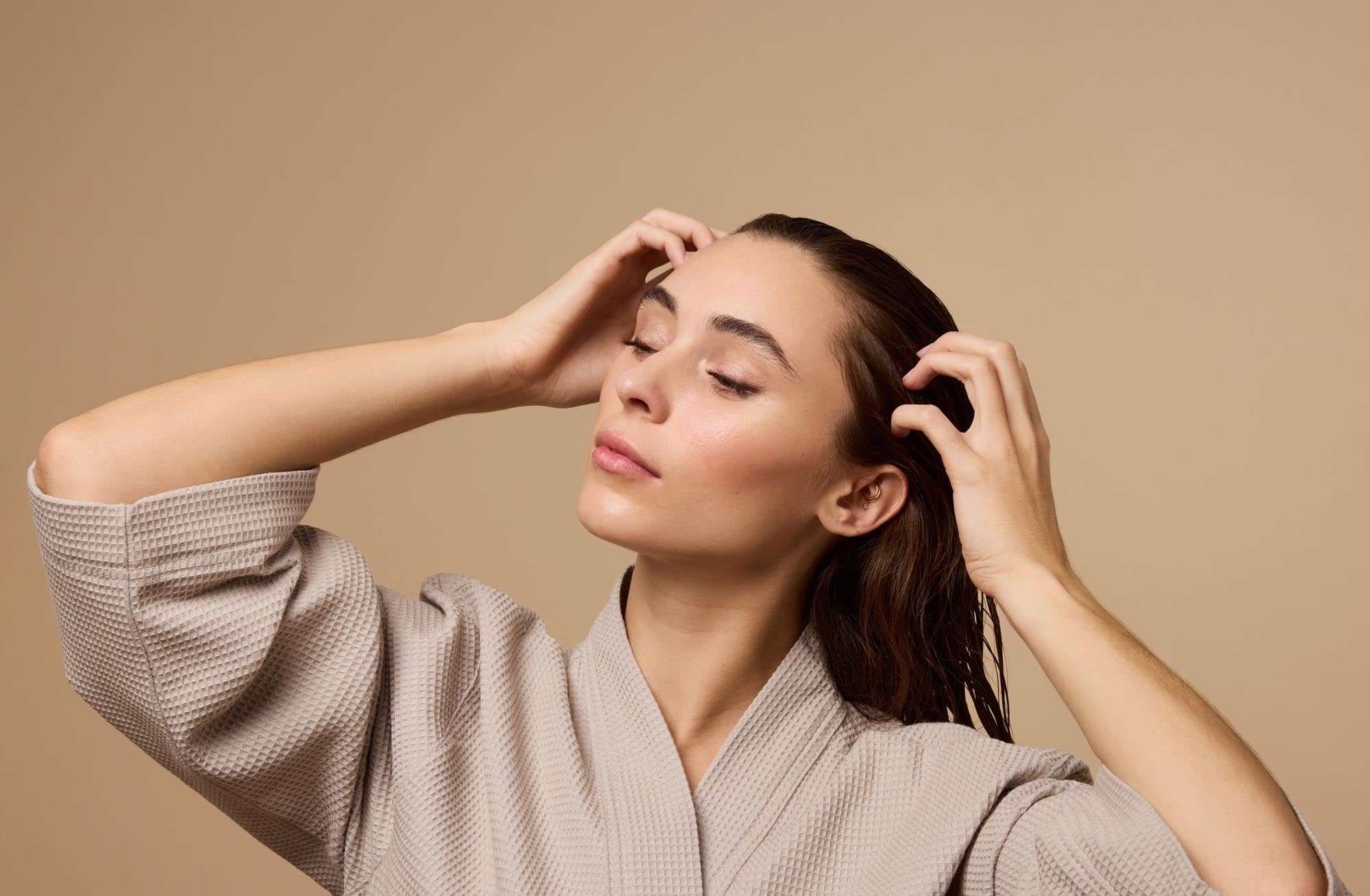 A woman with brown hair wears a robe and runs her fingers through her hair in front of a plain beige background.