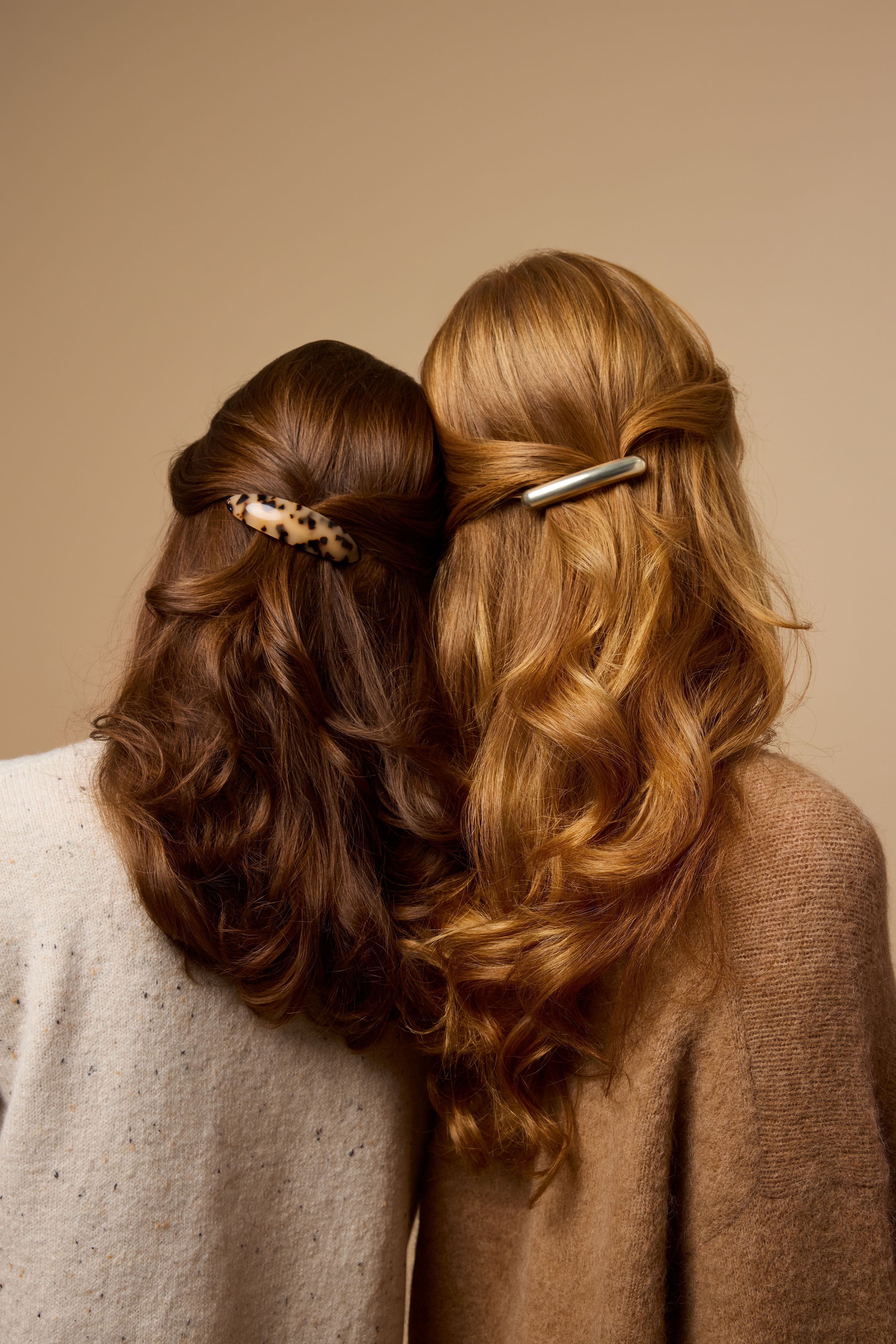 Two women with wavy hair, seen from behind, lean their heads together. One has brown hair with a tortoise-shell hair clip, and the other has blonde hair with a metallic hair clip. Both wear neutral-colored tops.