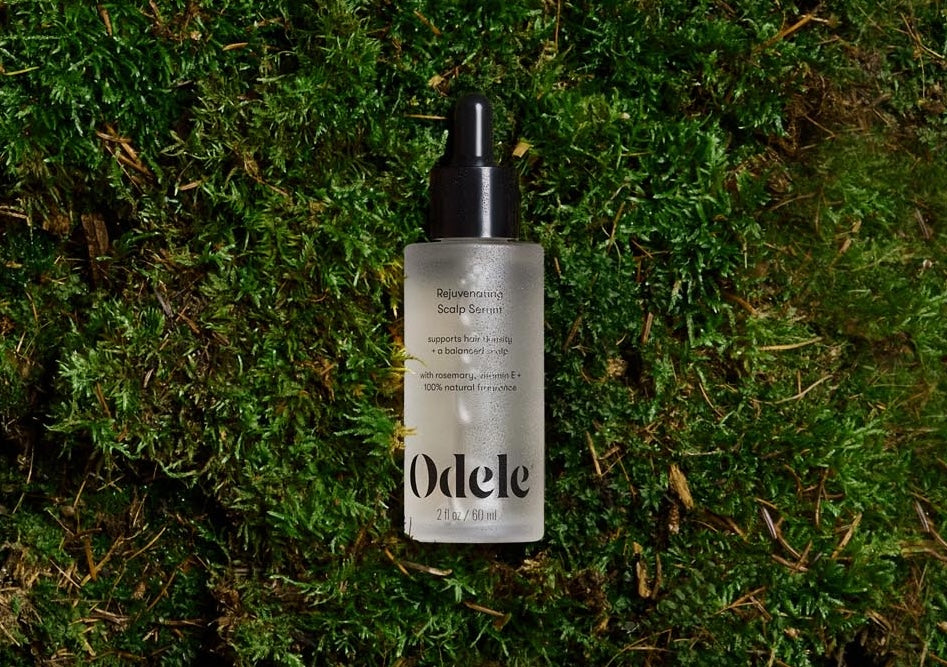 A bottle of Odele Rejuvenating Scalp Serum lays on a bed of green moss.