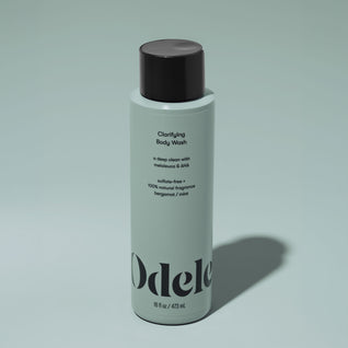 A bottle of Odele Clarifying Body Wash stands upright against a plain, light green backdrop with the front of the label facing the camera.