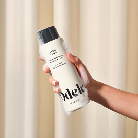A hand covered in soap suds holds a white bottle of Odele Beauty Clarifying Shampoo with a black cap, set against a softly blurred beige background.