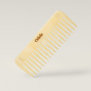 A light beige wide-toothed comb with a gold Odele logo on the handle stands at an angle against a plain, light-colored background.