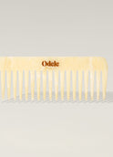 A light beige wide-toothed comb with a gold Odele logo on the handle stands upright against a plain, light-colored background.