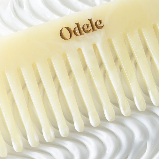 A light beige wide-toothed comb with a gold Odele logo on the handle lays on a background of swirling white cream.