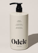 A beige bottle of Odele Leave-in Conditioner Bulk with a black pump top rests on a light surface. The label features detangling, plant proteins, 100% natural fragrance, and a 32 oz (946 ml) size.