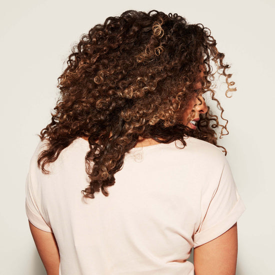 A woman with shoulder-length brown curls faces away from the camera in a light pink short-sleeve shirt against a plain off-white background.