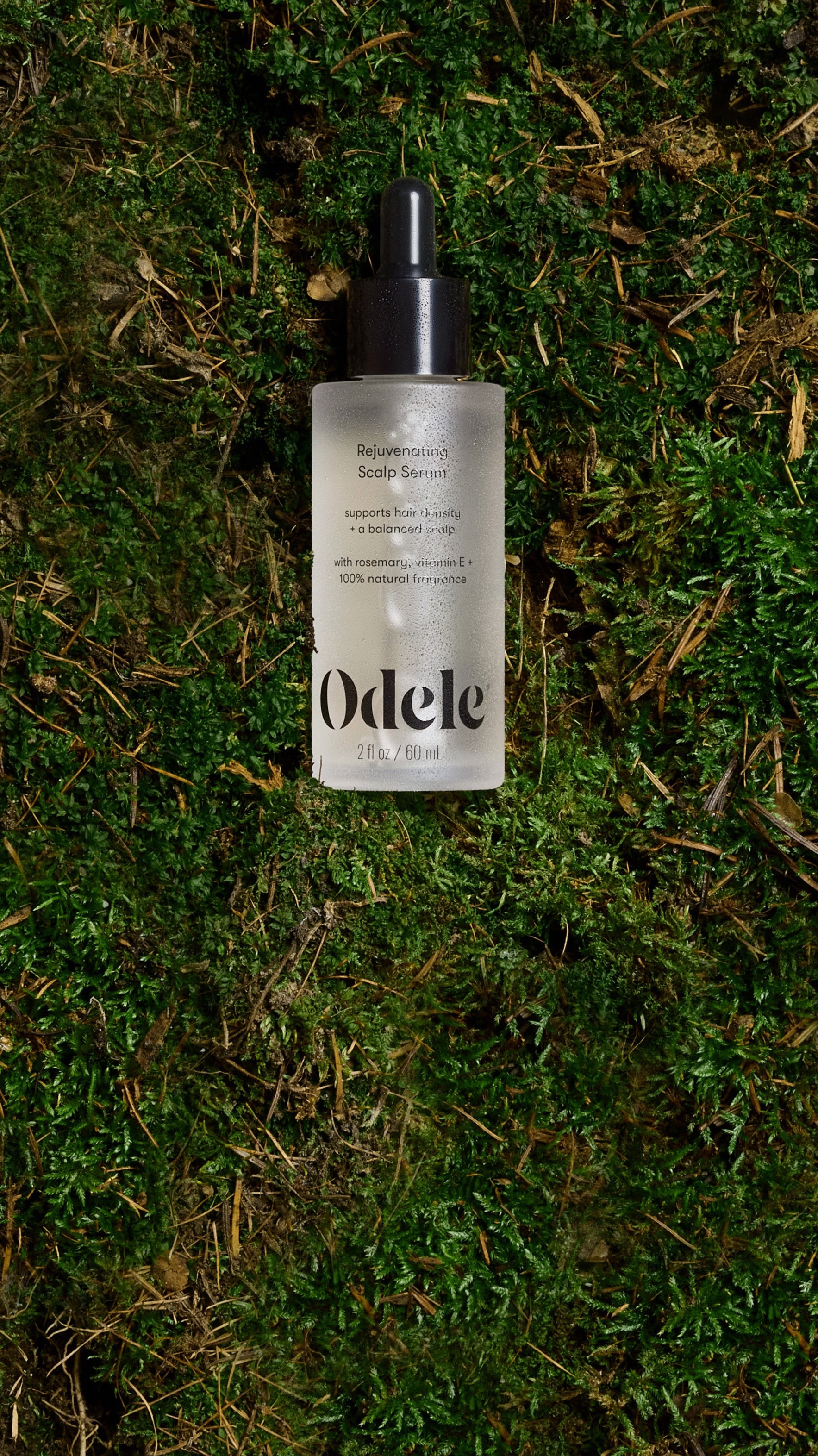 A bottle of Odele Rejuvenating Scalp Serum lays on a bed of green moss.