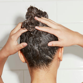 A woman with long dark hair applies Odele Scalp & Body Scrub to her hair in the shower.