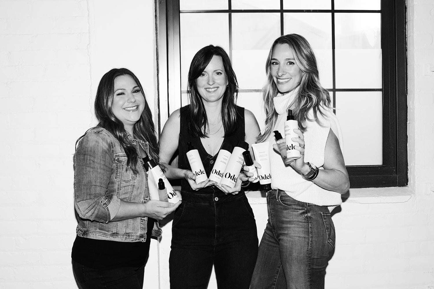 Three women stand side by side in front of a window, smiling and holding several skincare or beauty product bottles and tubes. The photo is in black and white.