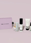 The Touch-Ups To Go Bundle is shown with a lavender Odele x Susan Alexandra gift box, beaded keychain, Hair Oil Mini, Air Dry Styler Mini, and a pump bottle of Odele hair care against a light pink background.