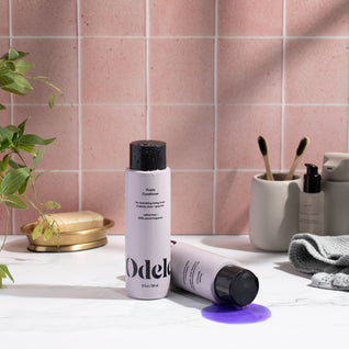 A bottle of Odele Purple Shampoo and Conditioner sitl on a white marble counter—one upright, one lying down with purple liquid spilled.