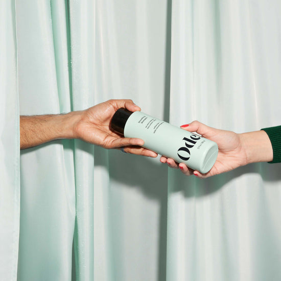 Two hands, one from each side, exchange a bottle of Odele Smoothing Shampoo in front of matching curtains.