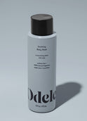 A bottle of Odele Soothing Body Wash stands upright against a plain light blue backdrop with the front of the label facing the camera.