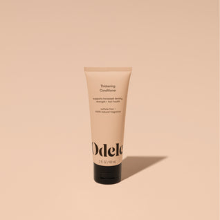 A tube of Odele Thickening Conditioner stands upright against a plain, light orange backdrop with the front of the label facing the camera.