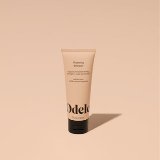 A tube of Odele Thickening Shampoo stands upright against a plain, light orange backdrop with the front of the label facing the camera.