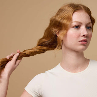 A young woman with long red wavy hair twists a section of her hair. She wears a short-sleeved white top and stands against a beige background, looking to the side.
