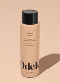 A bottle of Odele Thickening Shampoo stands upright against a plain, light orange backdrop with the front of the label facing the camera.