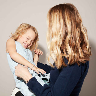 A woman with wavy blonde hair holds a laughing young girl wearing a light blue dress in front of a plain light-colored background.
