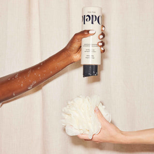 A soapy arm with medium brown skin squeezes Odele Ultra Sensitive Body Wash onto a white shower pouf held by another hand, with a neutral fabric background.