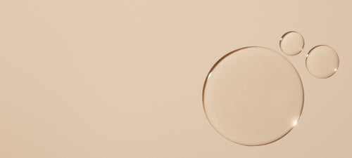 Three transparent liquid droplets rest on a smooth beige surface, with one large droplet on the right and two smaller droplets above it, creating a minimal and clean composition.