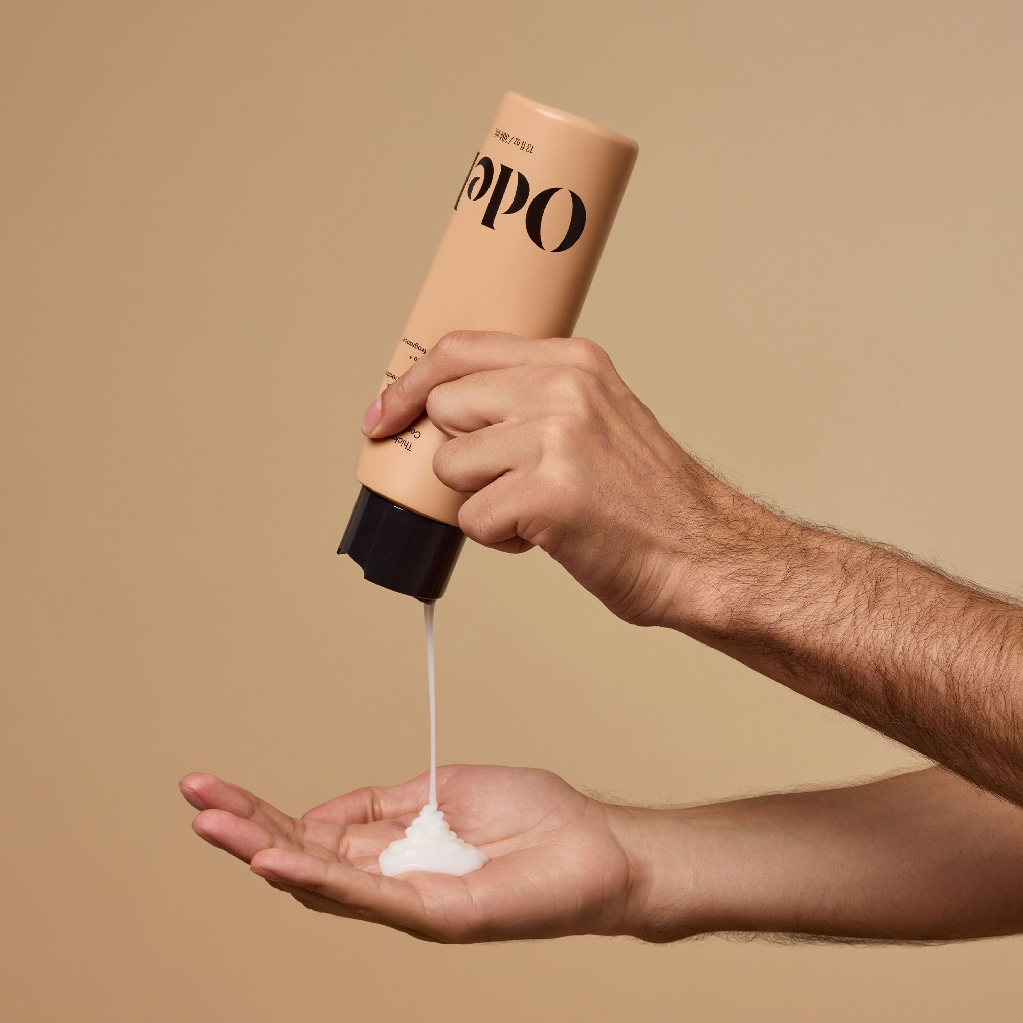 A hand squeezes a bottle of Odele Thickening Conditioner onto an open palm against a plain beige background.