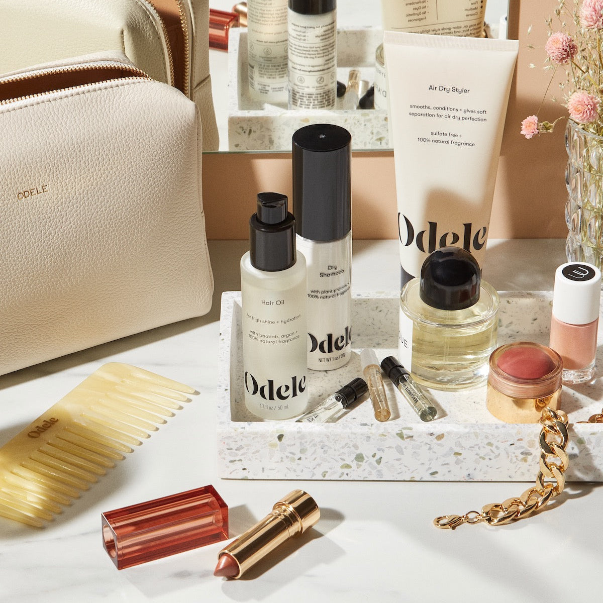 A collection of Odele products is arranged on a marble counter with bottles, lipstick, comb, and a gold bracelet beside a beige toiletry bag and pink flowers in the background.