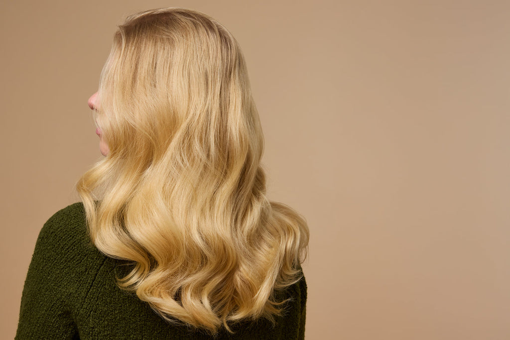 A person with long, thin wavy blonde hair is facing away, wearing a dark green textured top against a beige background.