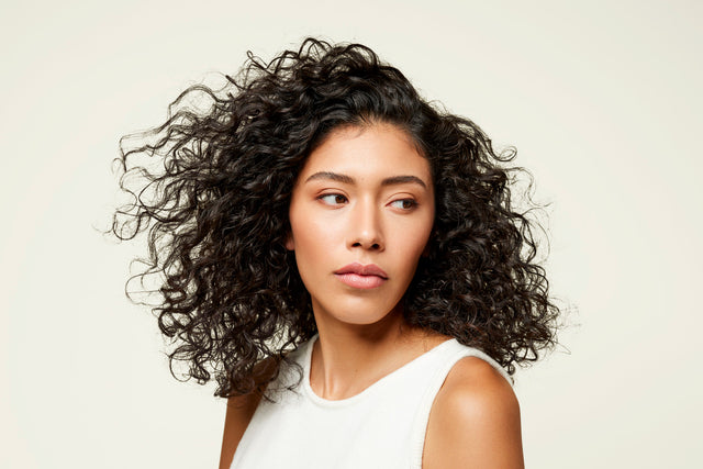 A person with dark hair and defined natural curls is seen wearing a white sleeveless top, looking to the side against a plain background.