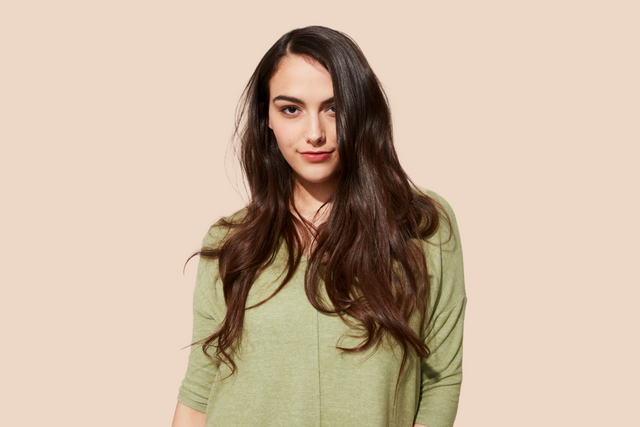 A person with shiny, smooth long brown hair wearing green pajamas stands against a plain beige background.
