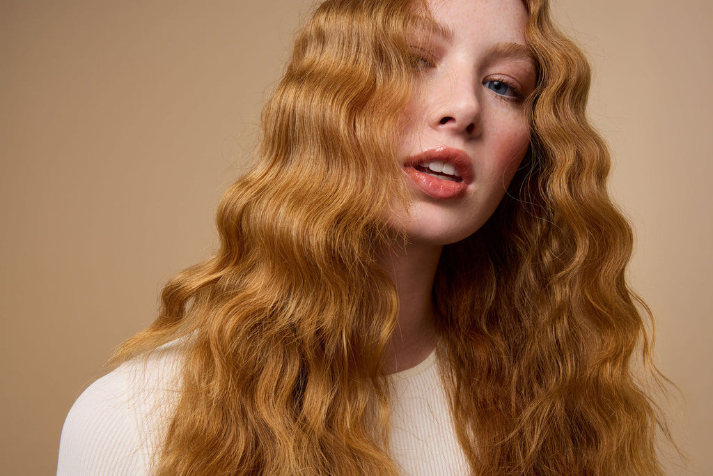 A woman with long, crimped red hair wearing a white shirt smiles softly at the camera.