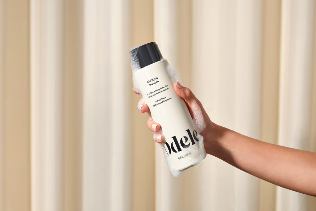 A hand holds a suds-covered bottle of Odele Clarifying Shampoo, a chelating shampoo, against a cream shower curtain backdrop.