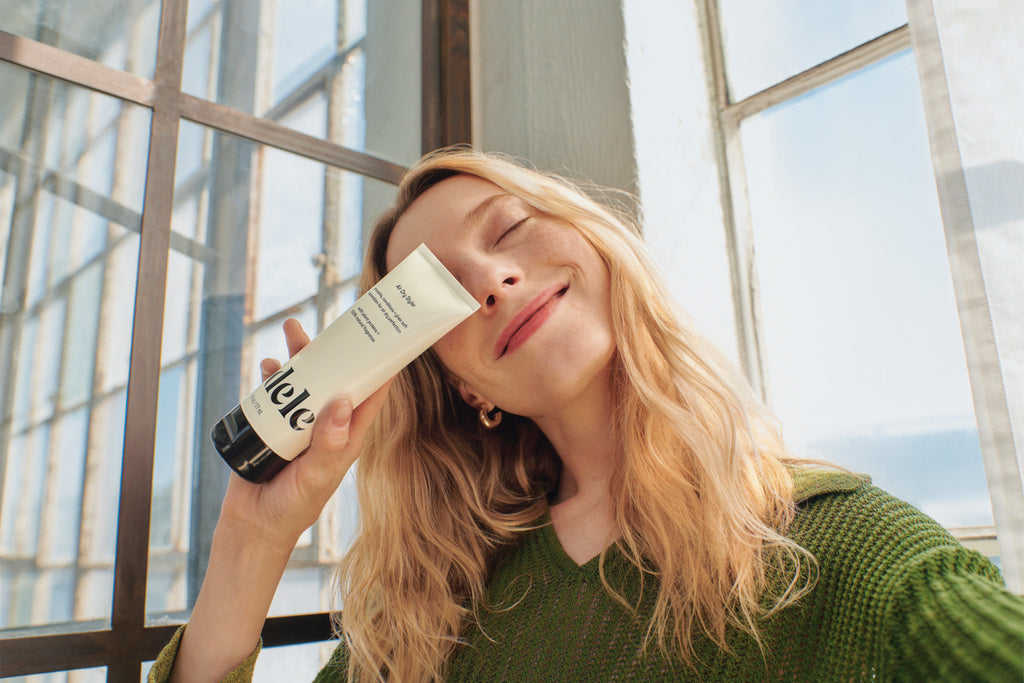 Woman with long, wavy blonde hair holds up a bottle of Odele Air Dry Styler.