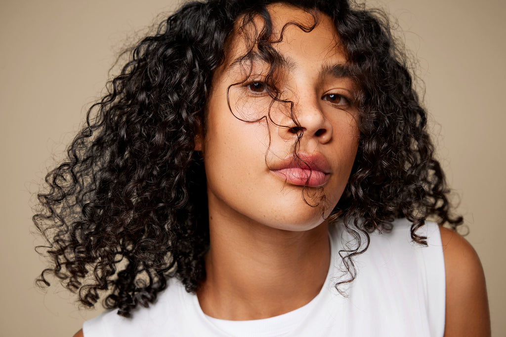 Person with curly, dark brown hair wearing a white shirt, looking directly at the camera with a neutral expression.