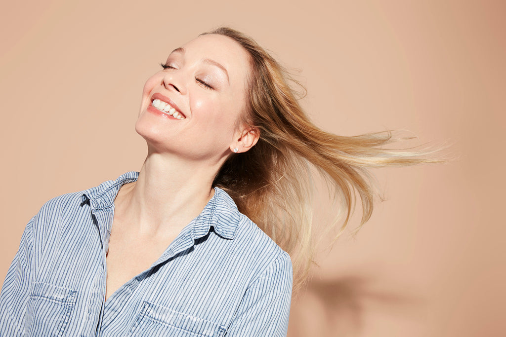 A woman with long, thin blonde hair smiles and tilts her head back, wearing a blue striped shirt against a beige background.