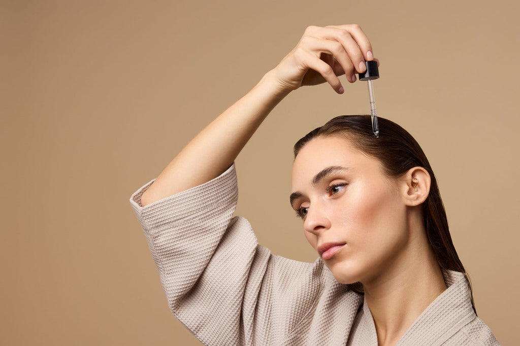 A person with damp brown hair wearing a robe applies Odele Rejuvenating Scalp Serum to their scalp.