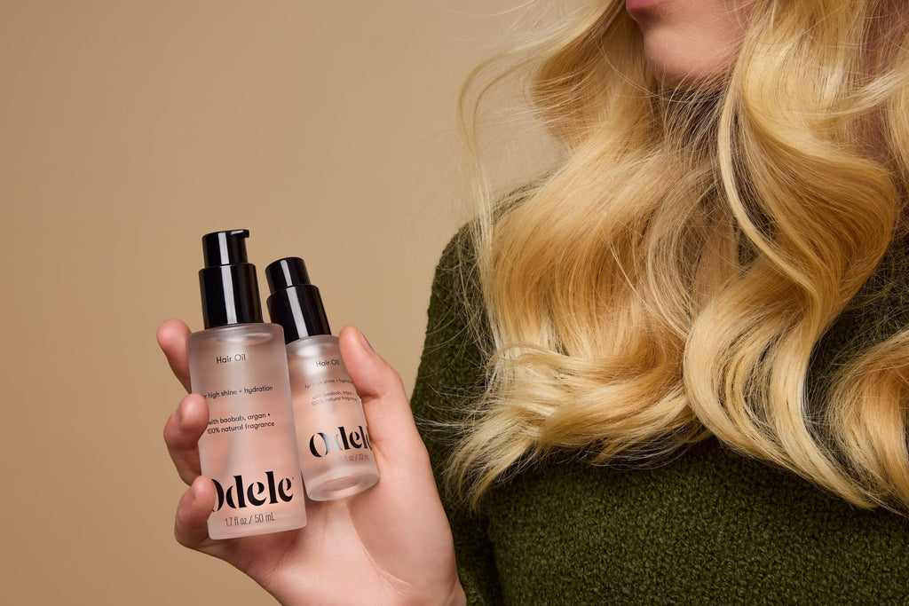 Woman with long blonde hair holding two bottles of Odele Hair Oil on a cream background