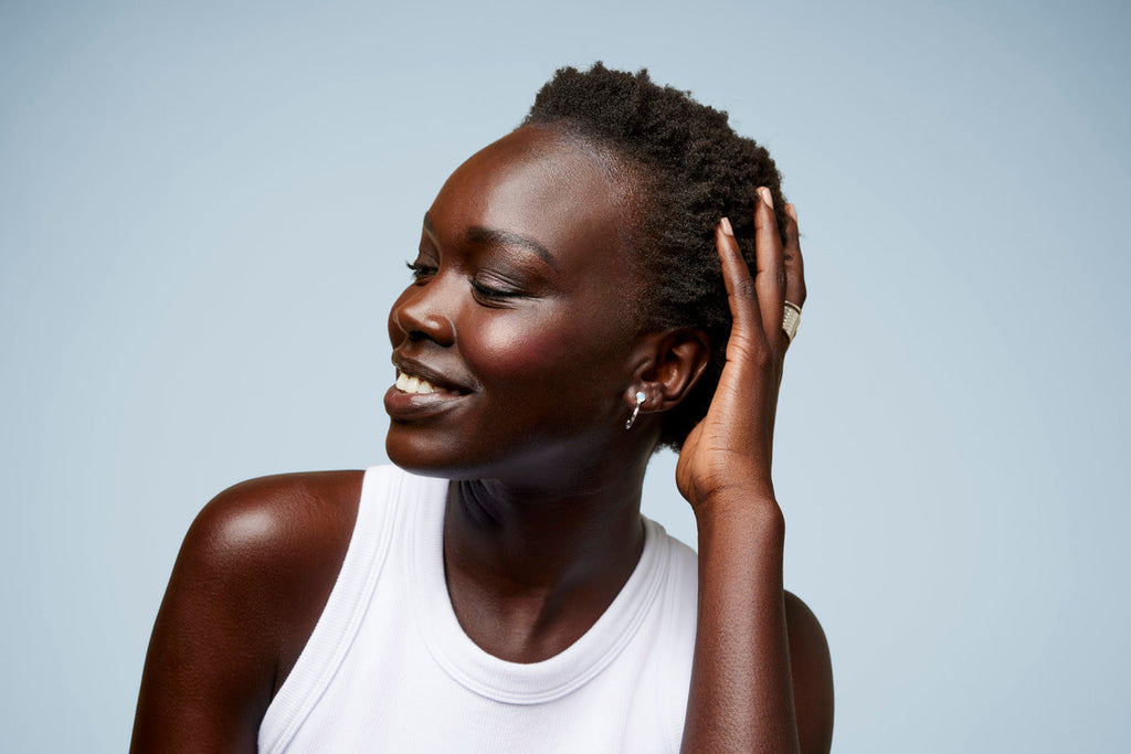 A person with short natural hair smiles while gently touching their head, wearing a white tank top against a blue background.