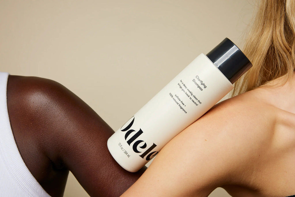 A person holds a white bottle of Odele Clarifying Shampoo against another person’s bare shoulder; both individuals have different skin tones.