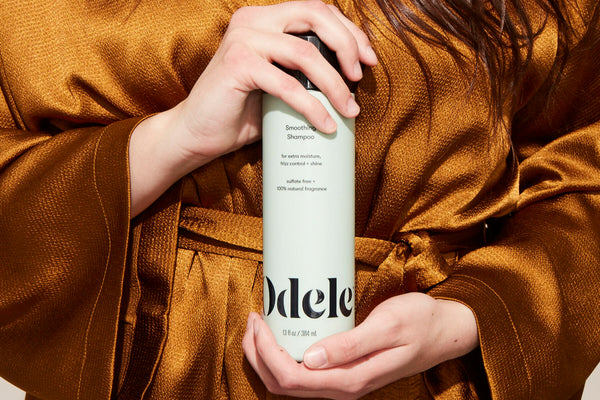 Woman wearing a silky copper-colored robe holds up a bottle of Odele Smoothing Shampoo
