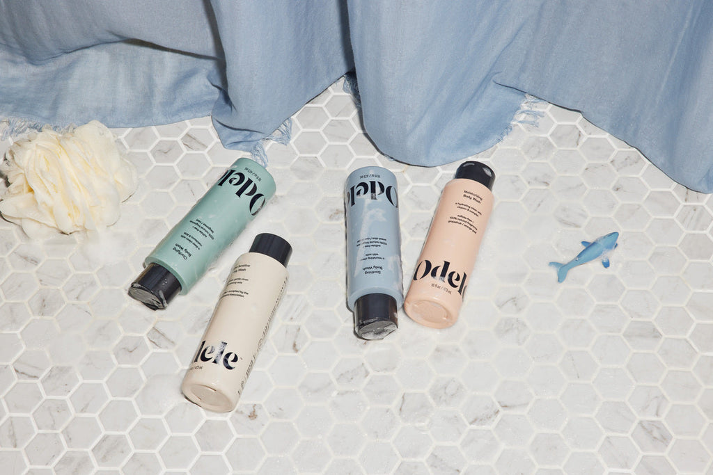 Flat lay of Odele body wash bottles on tile floor with a blue shower curtain backdrop, next to a cream shower pouf and small blue toy shark.