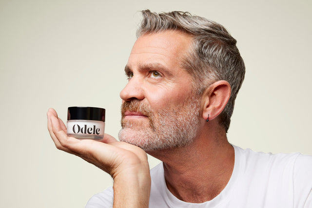 A middle-age man with sculpted gray hair and a neat beard holds up a jar of Odele Cream Pomade 