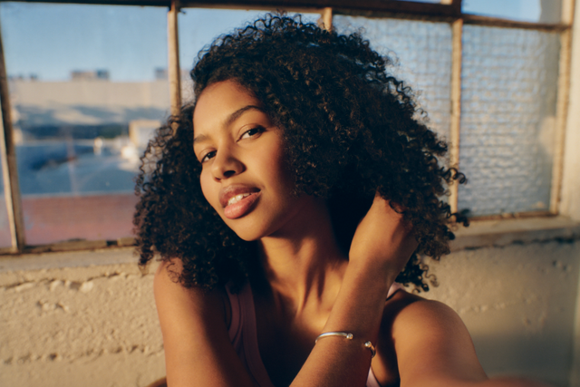 A person with natural curly hair poses for a selfie by the window as the golden hues of sunset frame their face.