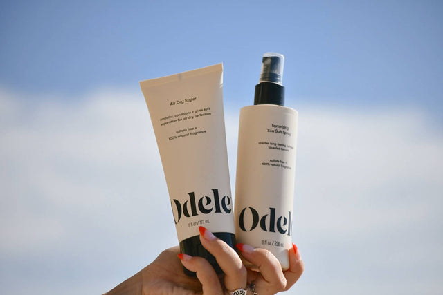A hand holds bottles of Odele Air Dry Styler and Texturizing Sea Salt Spray up to the sky 